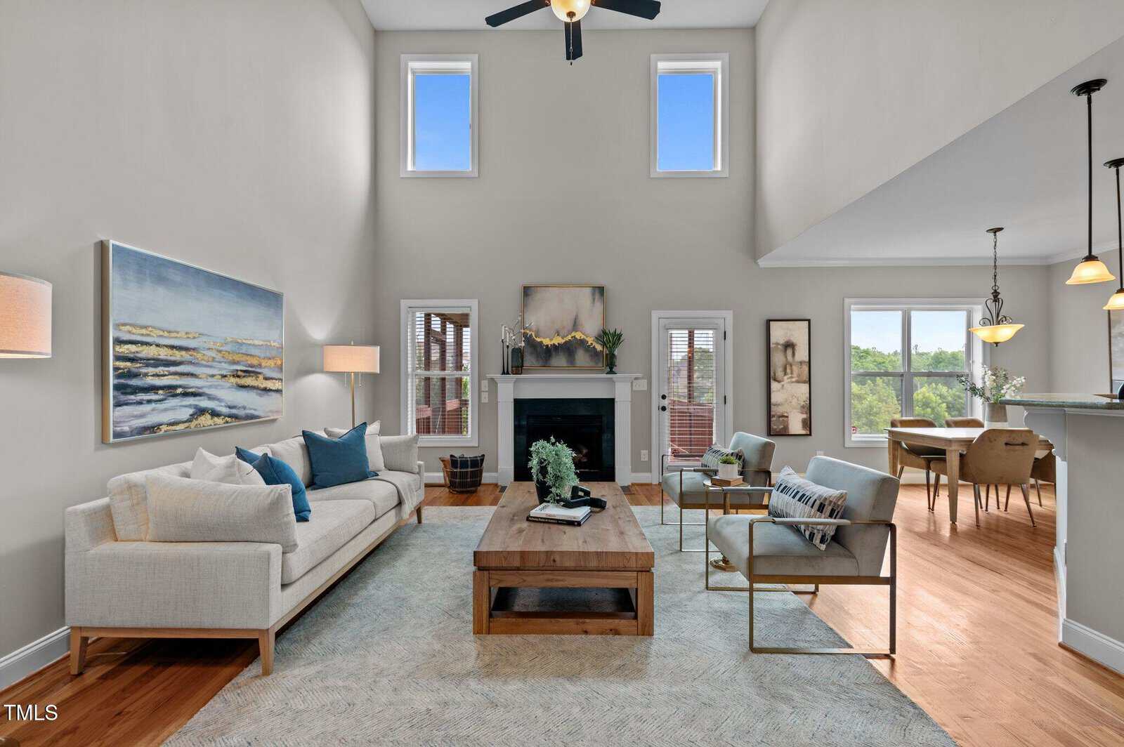 Bright living room with high ceiling, light walls, and wood floors. Features two sofas, a coffee table, fireplace, artwork, and large windows letting in natural light. Kitchen and dining area visible in the background.