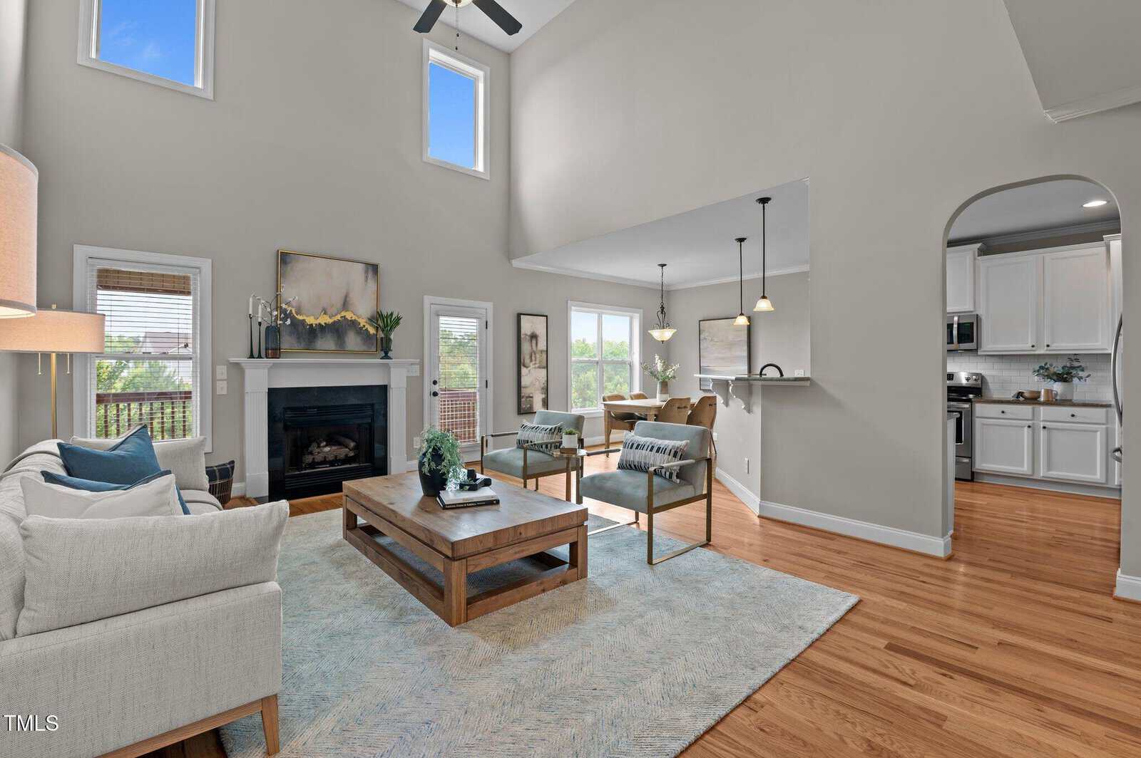 Bright living room with high ceilings, large windows, light gray walls, hardwood floors, a fireplace, and a cozy seating area. Adjacent dining area and modern white kitchen are visible through an arched doorway.