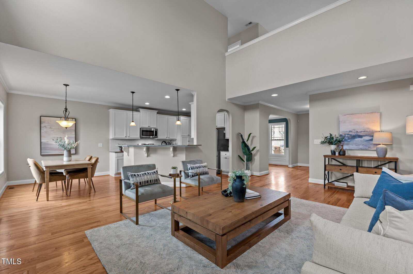 Modern open-plan living and dining area with light walls, wooden floors, a kitchen with white cabinets, a wooden dining table, and cozy seating around a wooden coffee table. Large windows bring in natural light.