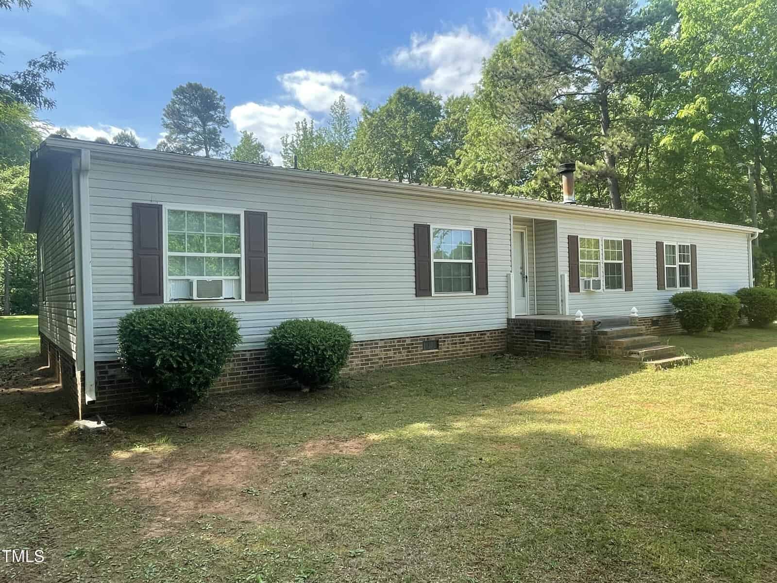 A single-story manufactured home at 6600 Clearwater Drive, Oxford, features white siding, brown shutters, and a brick foundation, surrounded by grass, shrubs, and trees under a partly cloudy sky.