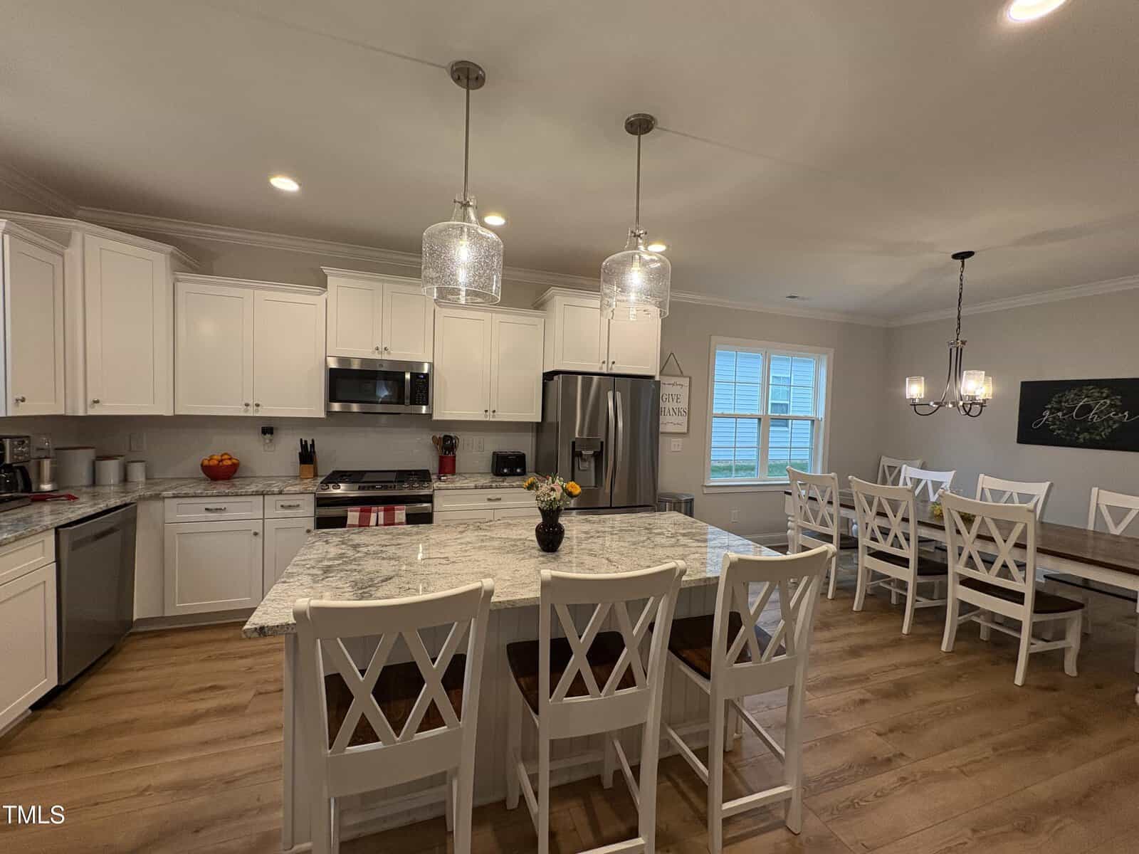 Spacious kitchen with white cabinets, stainless steel appliances, marble countertops, a large island with chairs, and a dining area with a long table and white chairs. Hardwood floors throughout and pendant lights above the island.