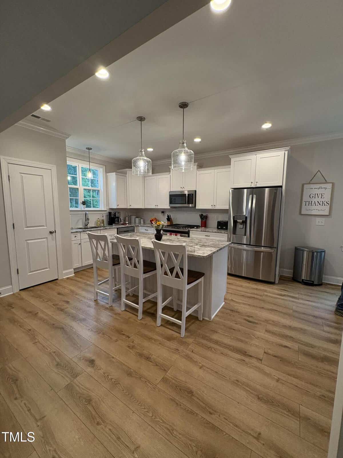 Modern kitchen with white cabinets, stainless steel appliances, marble island with three chairs, wood flooring, two pendant lights above island, window above sink, and a “Give Thanks” wall sign near a trash can.