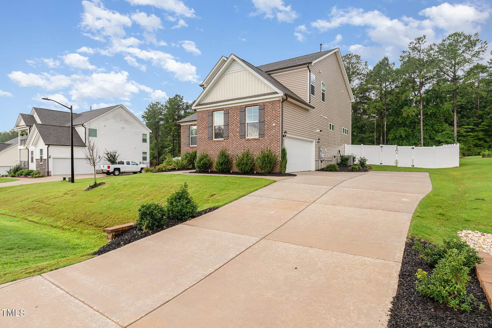 Two-story beige house with a white garage door, a sloped driveway, green lawn, and landscaping. Another house and a white truck are visible in the background under a partly cloudy sky.