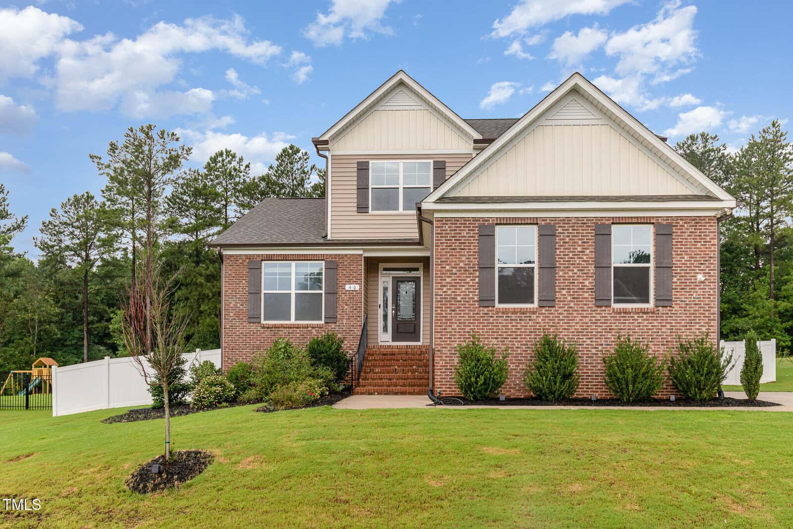 Two-story brick and siding house located at 40 Ironwood Boulevard in Youngsville, featuring front steps, shrubs, a manicured lawn, white fence, and trees in the background under a partly cloudy sky.