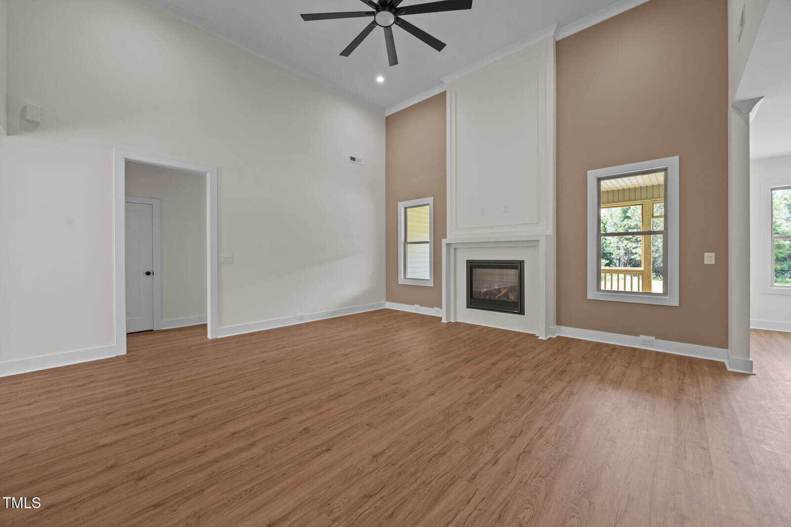 Spacious empty living room with wood floors, a ceiling fan, a tall white-trimmed fireplace, beige accent walls, and two windows flanking the fireplace. Doorways lead to adjacent rooms and a patio is visible through a window.