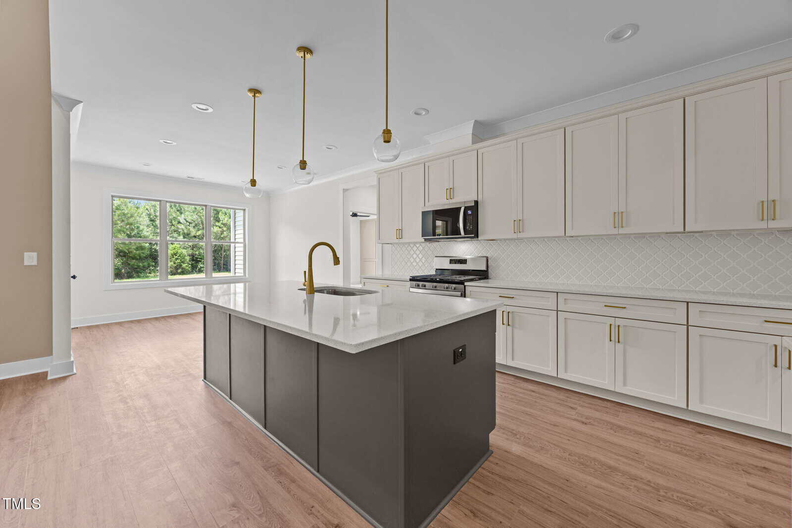 Modern kitchen with a large island, gold pendant lights, white cabinets, stainless steel appliances, a geometric tile backsplash, wood flooring, and a large window letting in natural light.