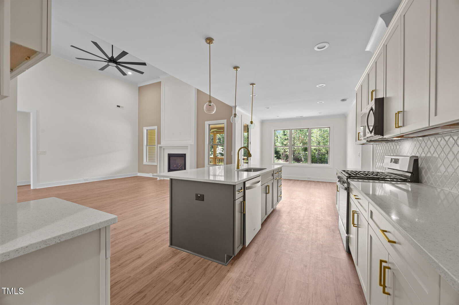 Modern kitchen with white cabinets, gold hardware, a center island, stainless steel appliances, and pendant lights. Open layout leads to a living area with a fireplace and large windows. Light wood floors throughout.