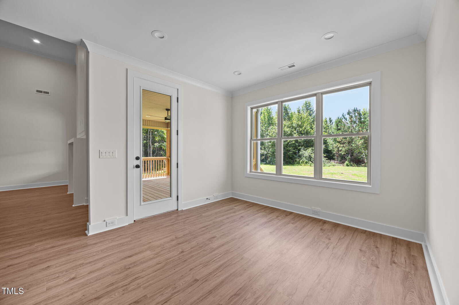 A bright, empty room with light-colored walls, wood-style flooring, large windows showing greenery outside, and a glass door leading to a covered porch. Ceiling lights are recessed. The space is clean and unfurnished.
