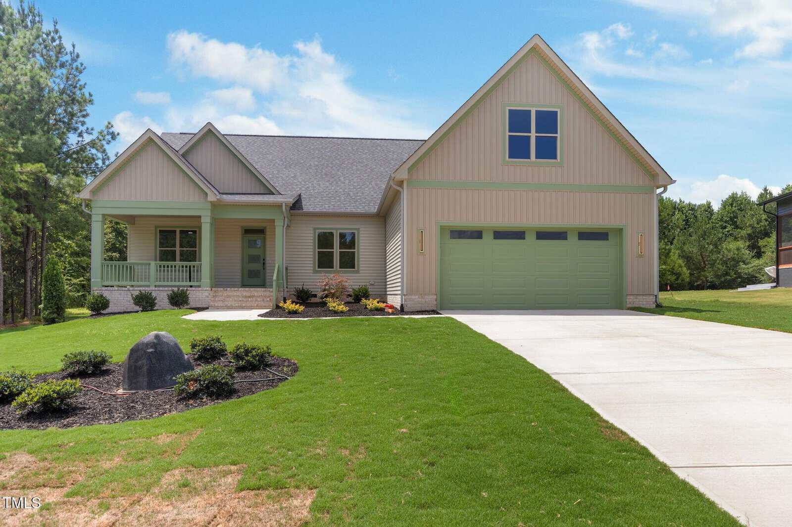 A modern single-story house with light green siding, a two-car garage, and a covered front porch. The yard is landscaped with shrubs and a rock, and a driveway leads to the garage. Pine trees and blue sky are in the background.