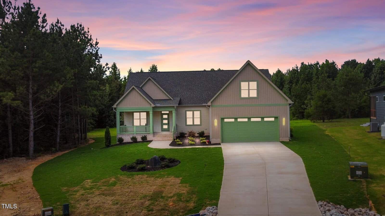 A light green, single-story house with a covered front porch and a three-car garage, sits on a large grassy lawn with trees in the background, under a colorful sunset sky.