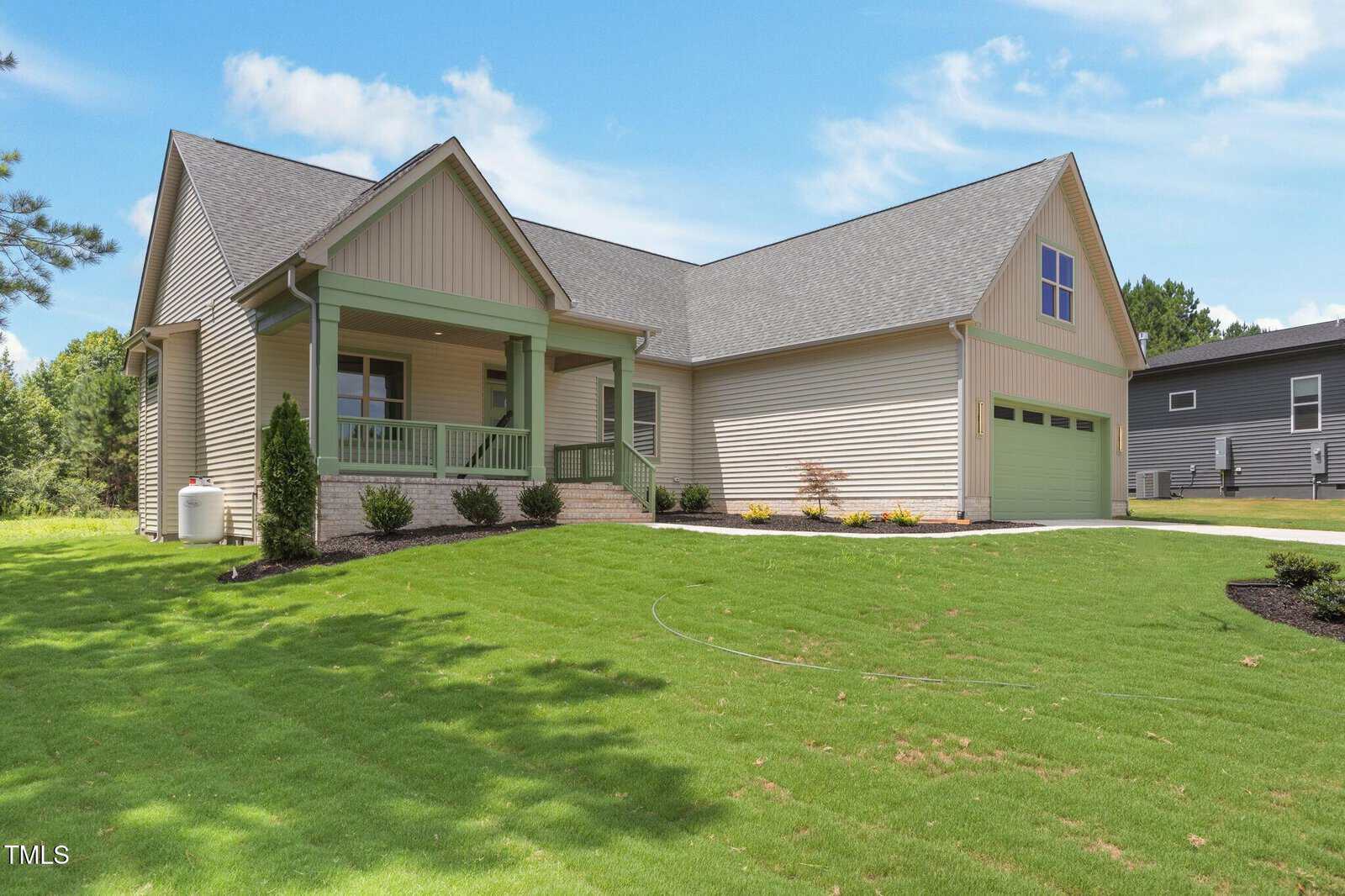 A modern single-story beige house with a covered front porch, green garage doors, neatly landscaped yard, and a bright green lawn under a blue sky with scattered clouds.
