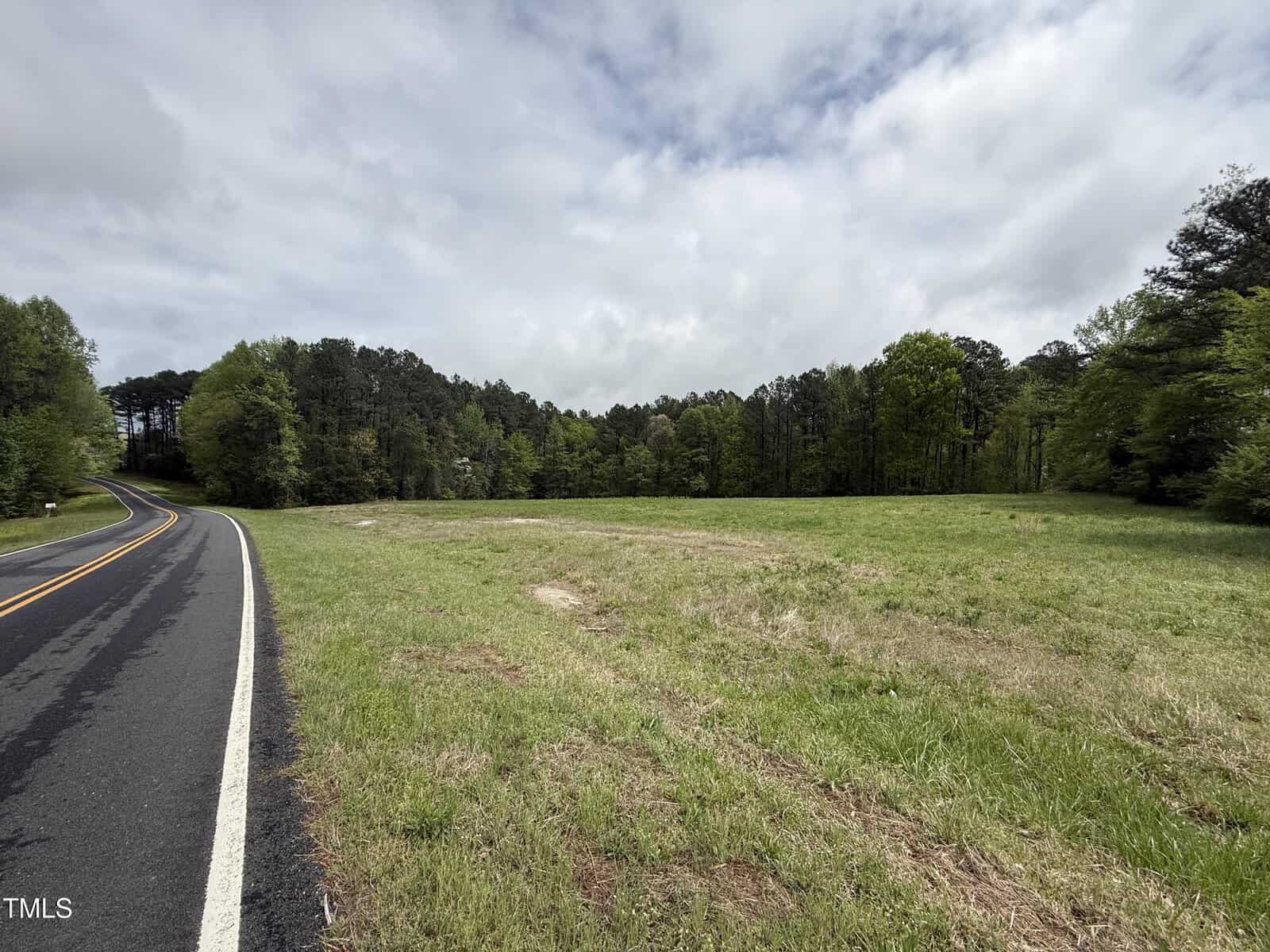 A paved two-lane road curves next to a grassy field bordered by dense trees under a partly cloudy sky. The scene, located on Stallings Mill Road near Louisburg, appears rural and calm, with no vehicles or people visible.