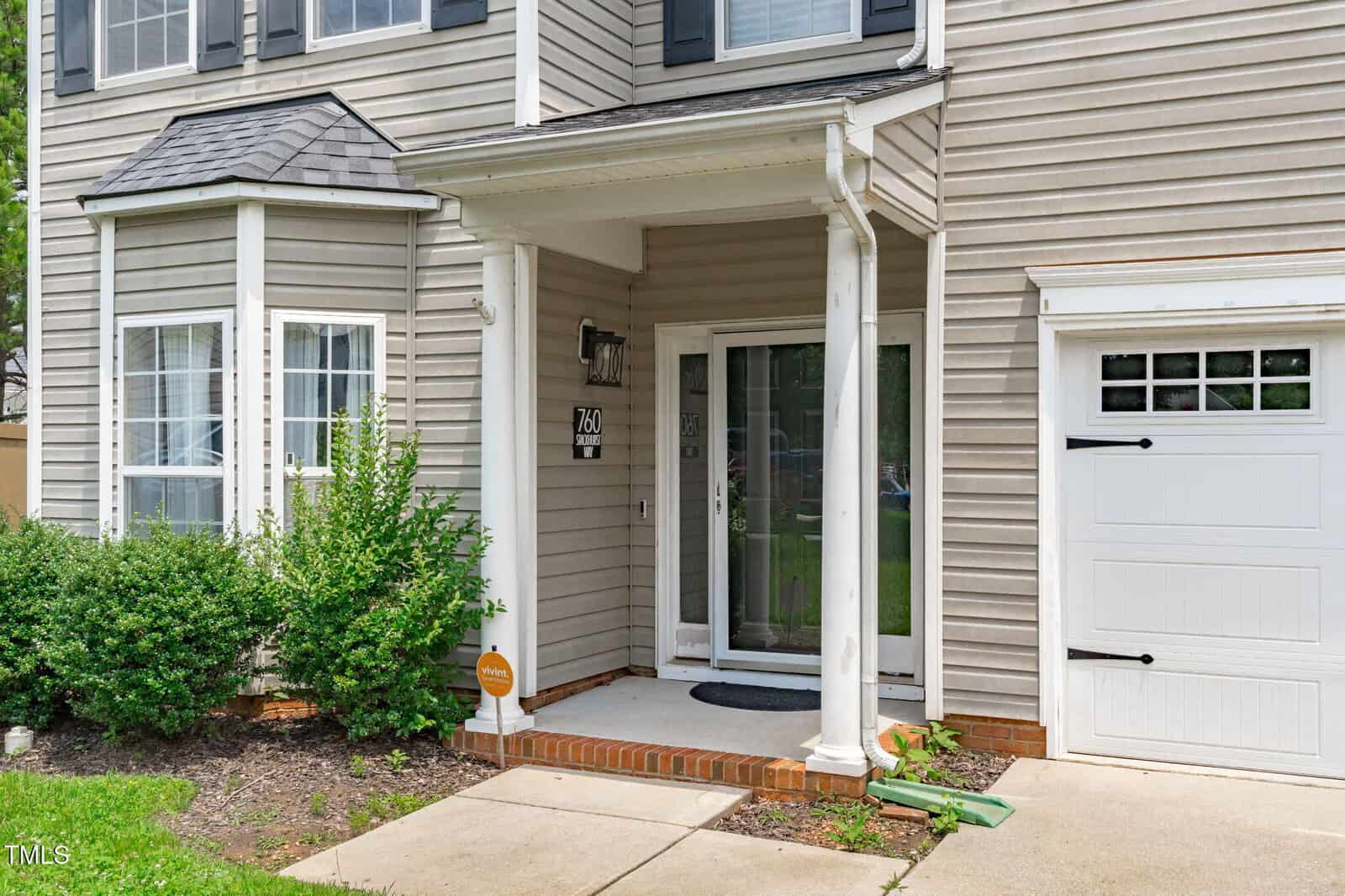 Front entrance of a beige house with white trim, glass front door, and small covered porch. Bushes and plants line the left side, and a white garage door is visible on the right. A sidewalk leads to the porch steps.