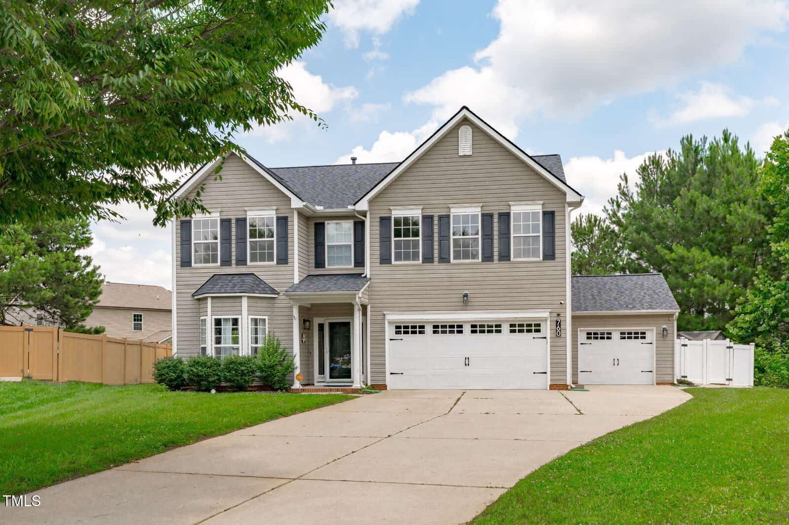 Two-story beige suburban house with white trim, black shutters, and a three-car garage. The house has a large driveway, green lawn, and is surrounded by trees and a wooden fence under a partly cloudy sky.