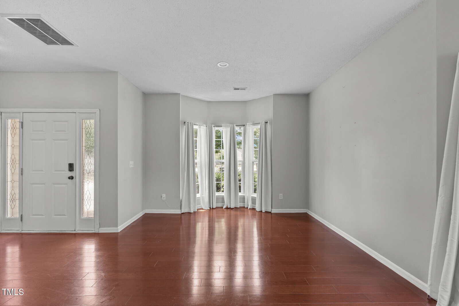 Empty room with red-brown hardwood floors, light gray walls, and a bay window with white curtains. The front door is on the left side, and the ceiling has a vent and light fixture. The space is well-lit by natural light.