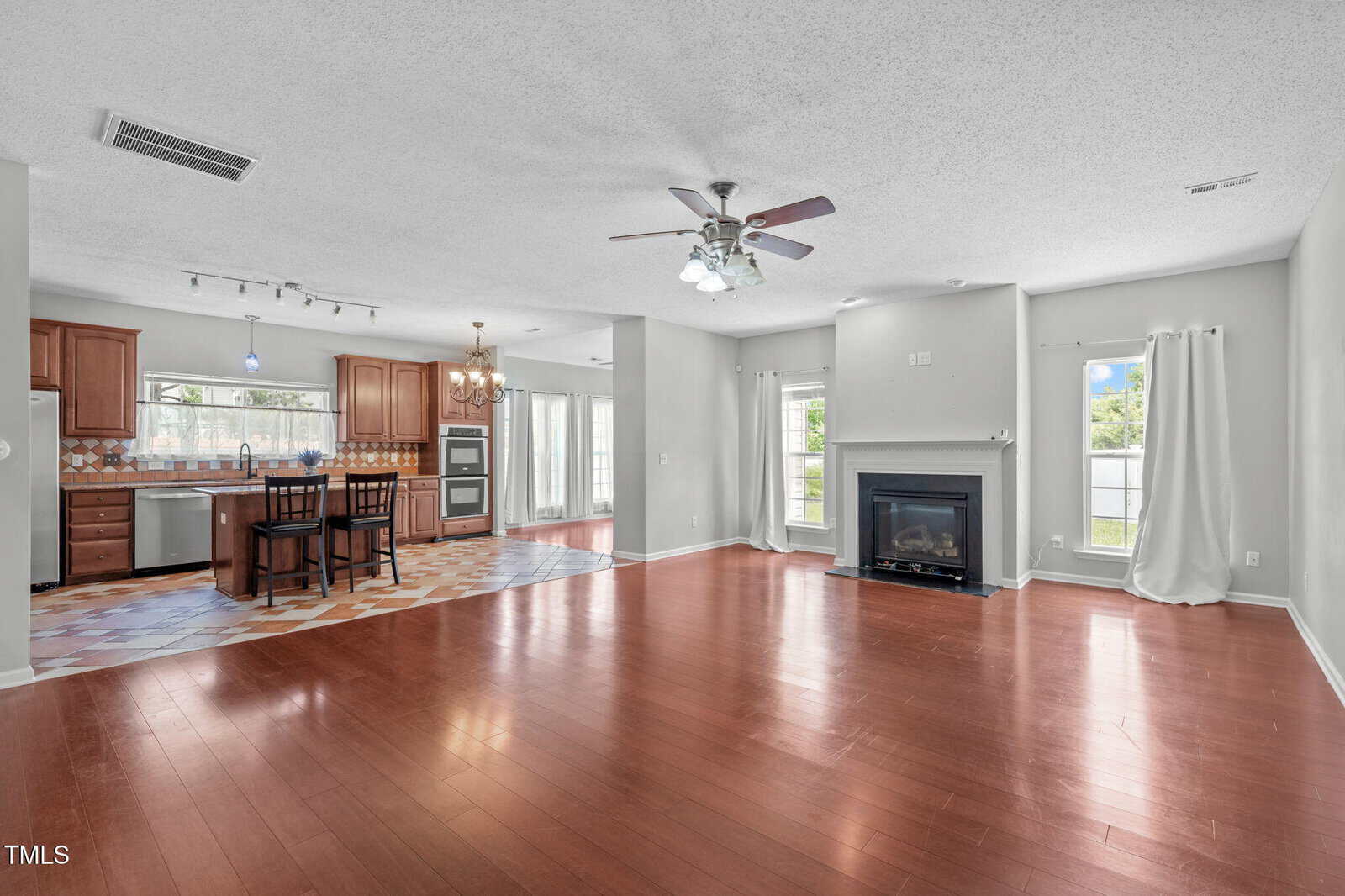 Open-concept living area with hardwood floors, a ceiling fan, a central fireplace, large windows with white curtains, and an adjoining kitchen with wooden cabinets, island, and tile flooring.