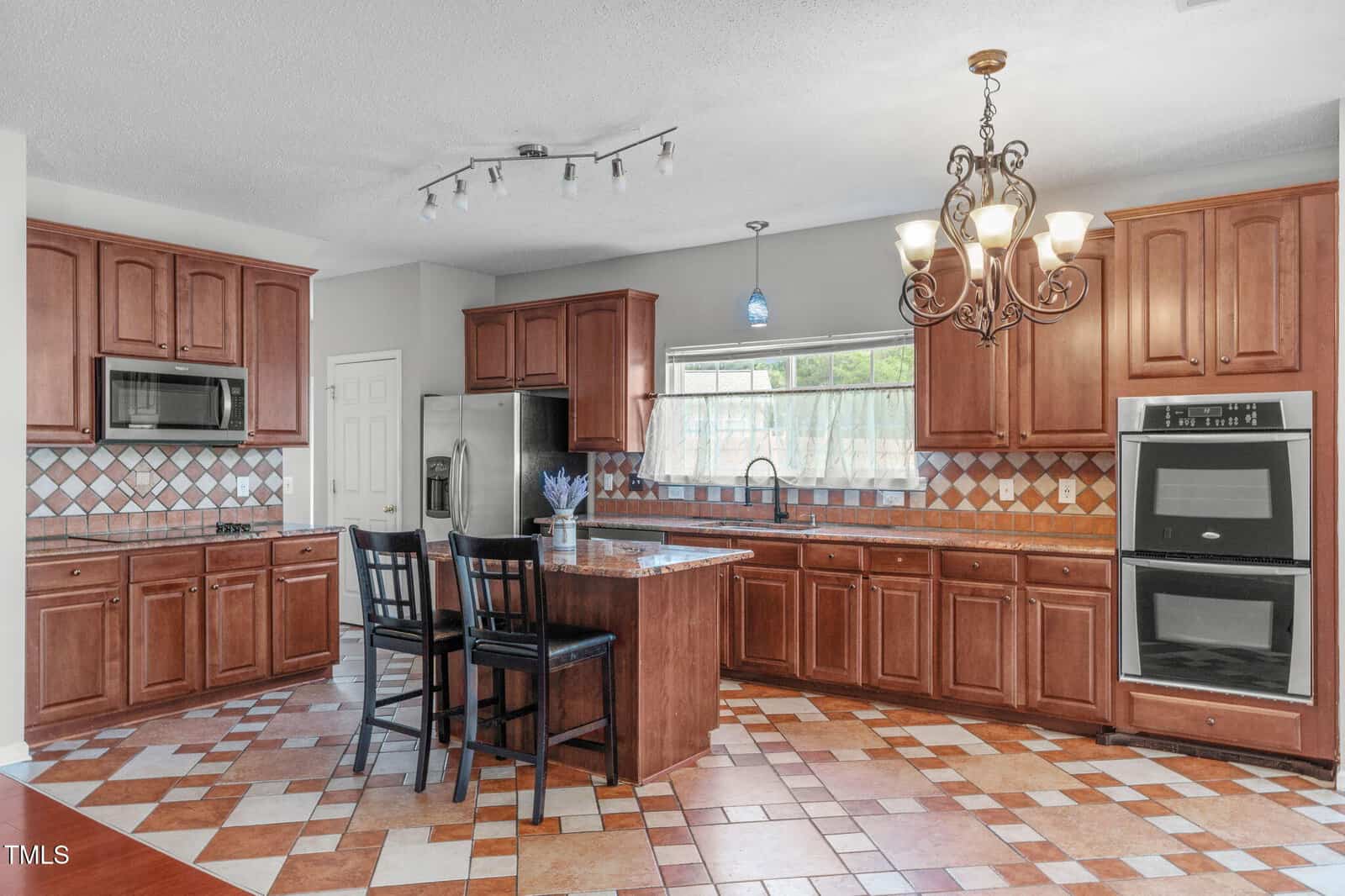 Spacious kitchen with brown cabinets, stainless steel appliances, a center island with two barstools, double oven, tiled backsplash, and multicolored tiled floor. A chandelier and track lighting provide illumination.
