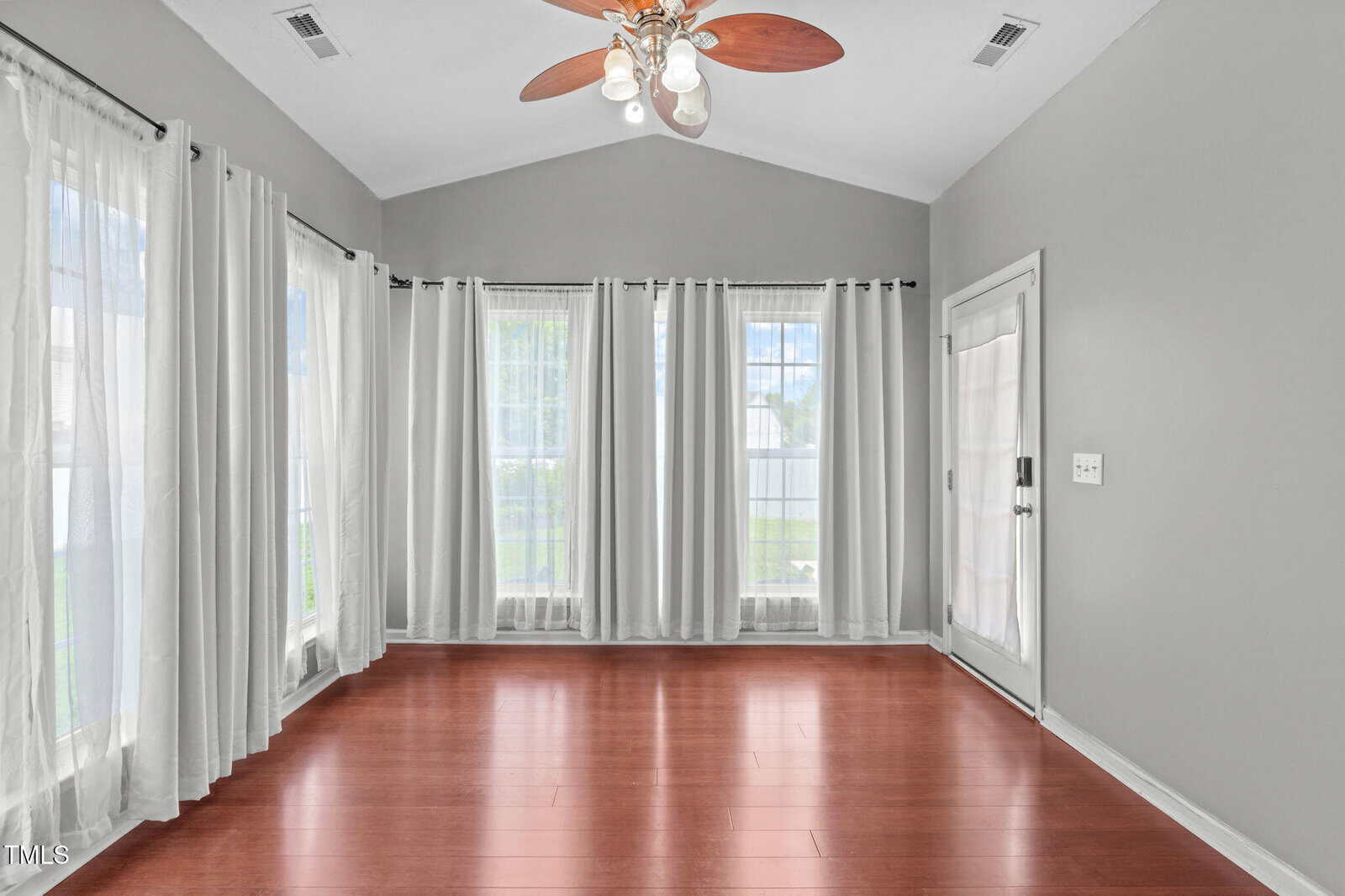 A bright, empty room with light gray walls, wood floors, large windows with sheer white curtains, a glass door, and a ceiling fan with lights. Natural light fills the space.