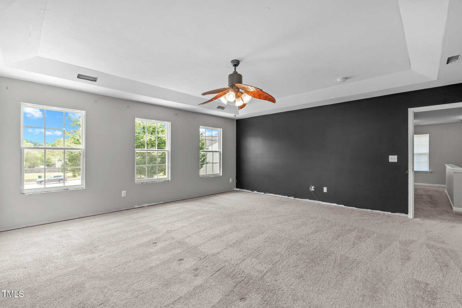 A spacious, empty room with light gray walls, a dark accent wall, large windows letting in natural light, a ceiling fan with wooden blades, and beige carpet flooring. An open doorway leads to another area.