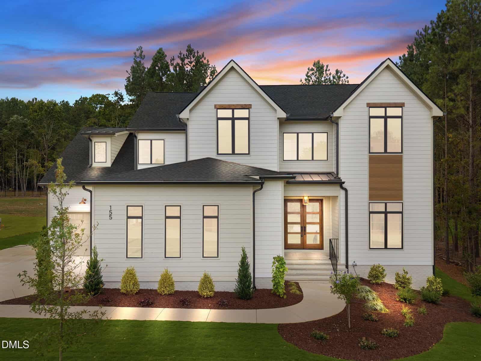 Modern two-story house with white siding, large windows, and a black roof at sunset, located at 155 Cherry Bark Drive in Youngsville. The front yard is landscaped with small shrubs, a curved driveway, and a few young trees, set against a backdrop of tall trees.