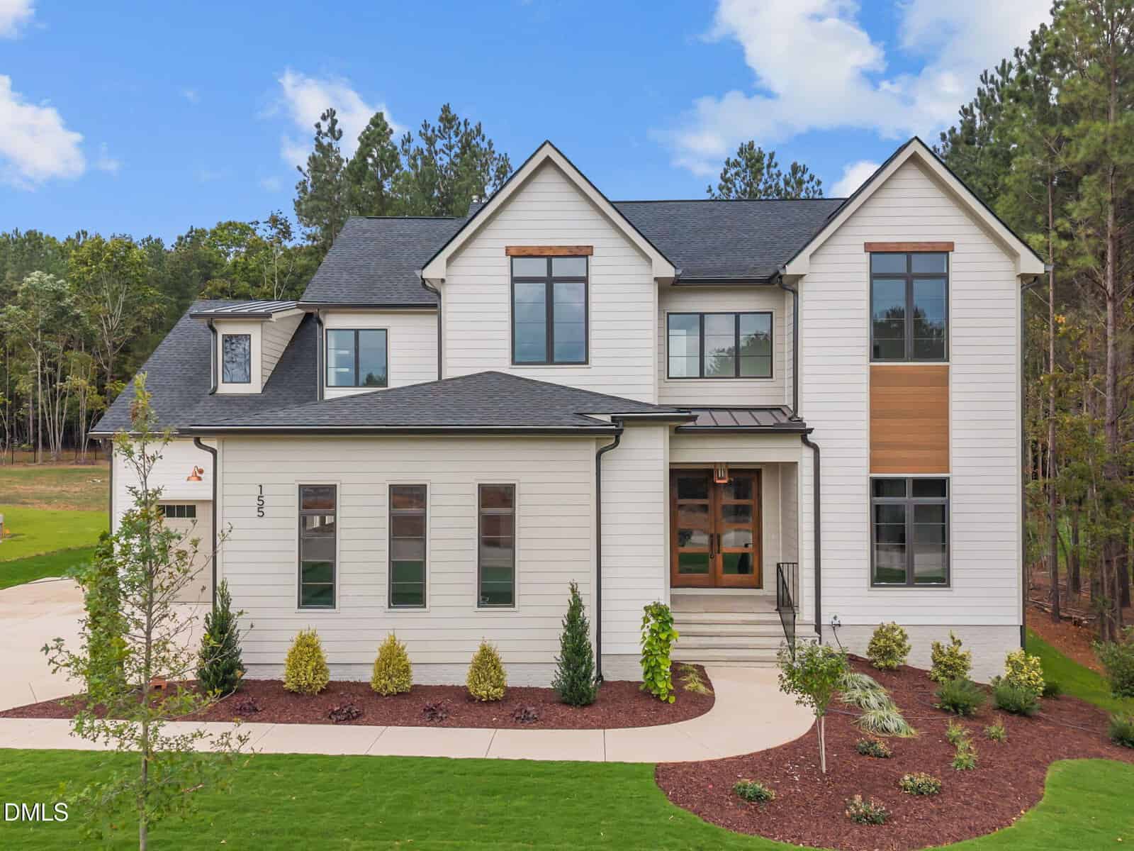 Two-story modern house with white siding, large windows, black roof, a landscaped front yard, and concrete walkway. Surrounded by trees and blue sky with scattered clouds in the background.