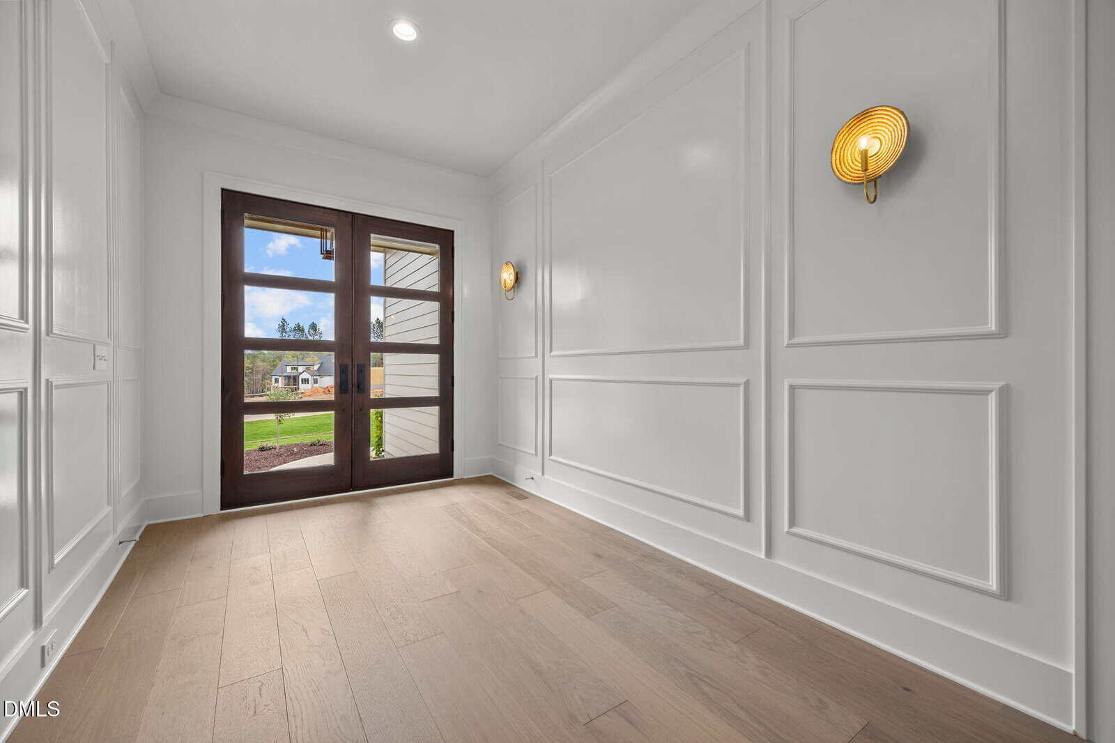 A bright entryway with light wood flooring, white paneled walls, two round gold wall sconces, and large glass double doors overlooking a green lawn and houses outside.