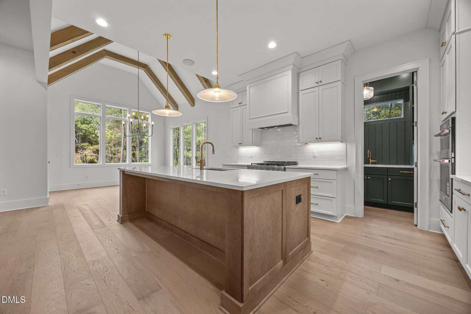 Modern kitchen with light wood floors, a large island with a white countertop, white cabinets, gold fixtures, and pendant lights. Exposed beams on the ceiling and large windows provide natural light. A pantry is visible to the right.
