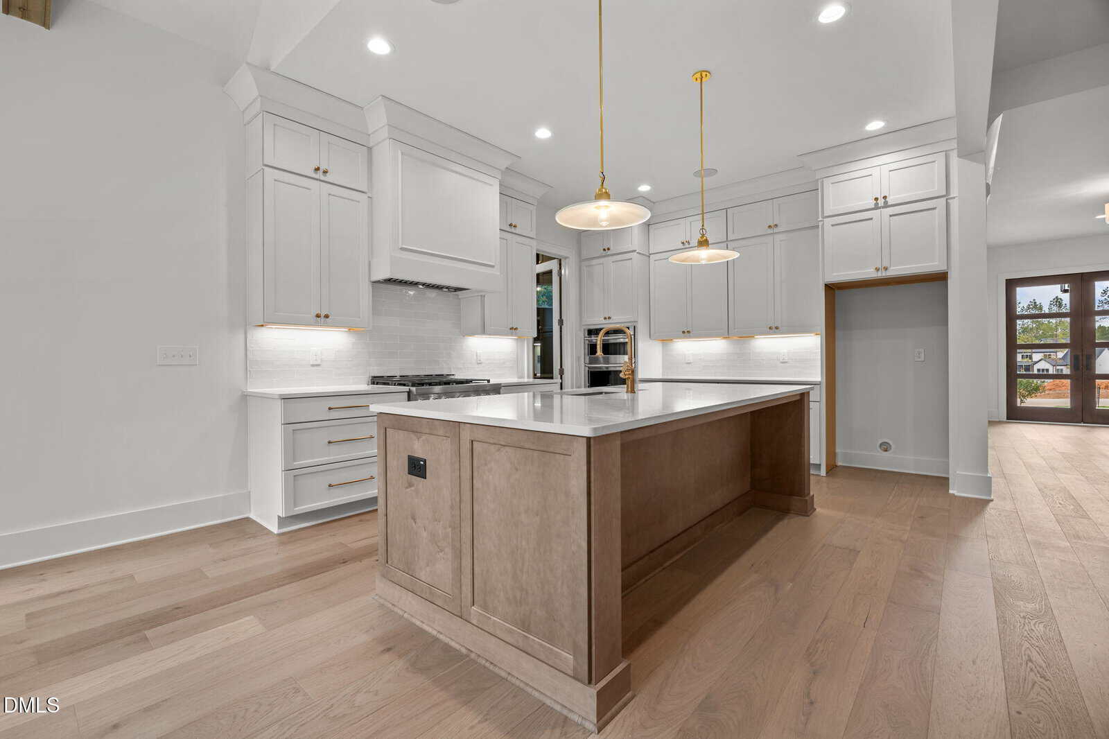 Modern kitchen with white cabinetry, stainless steel appliances, a large island with a light wood base, pendant lighting, and light wood flooring. White walls and ceiling with recessed lighting. Doorway visible in the background.