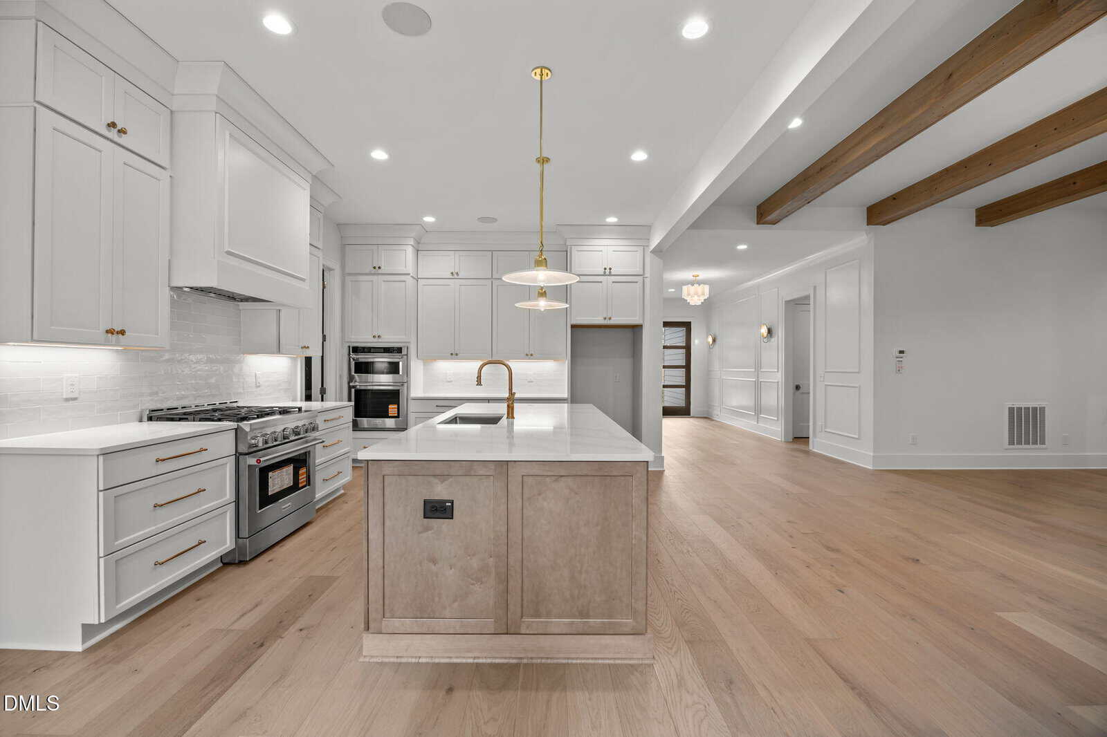 Modern kitchen with white cabinets, stainless steel appliances, a center island with a sink, pendant lighting, light wood flooring, and wooden ceiling beams, leading into an open living area.