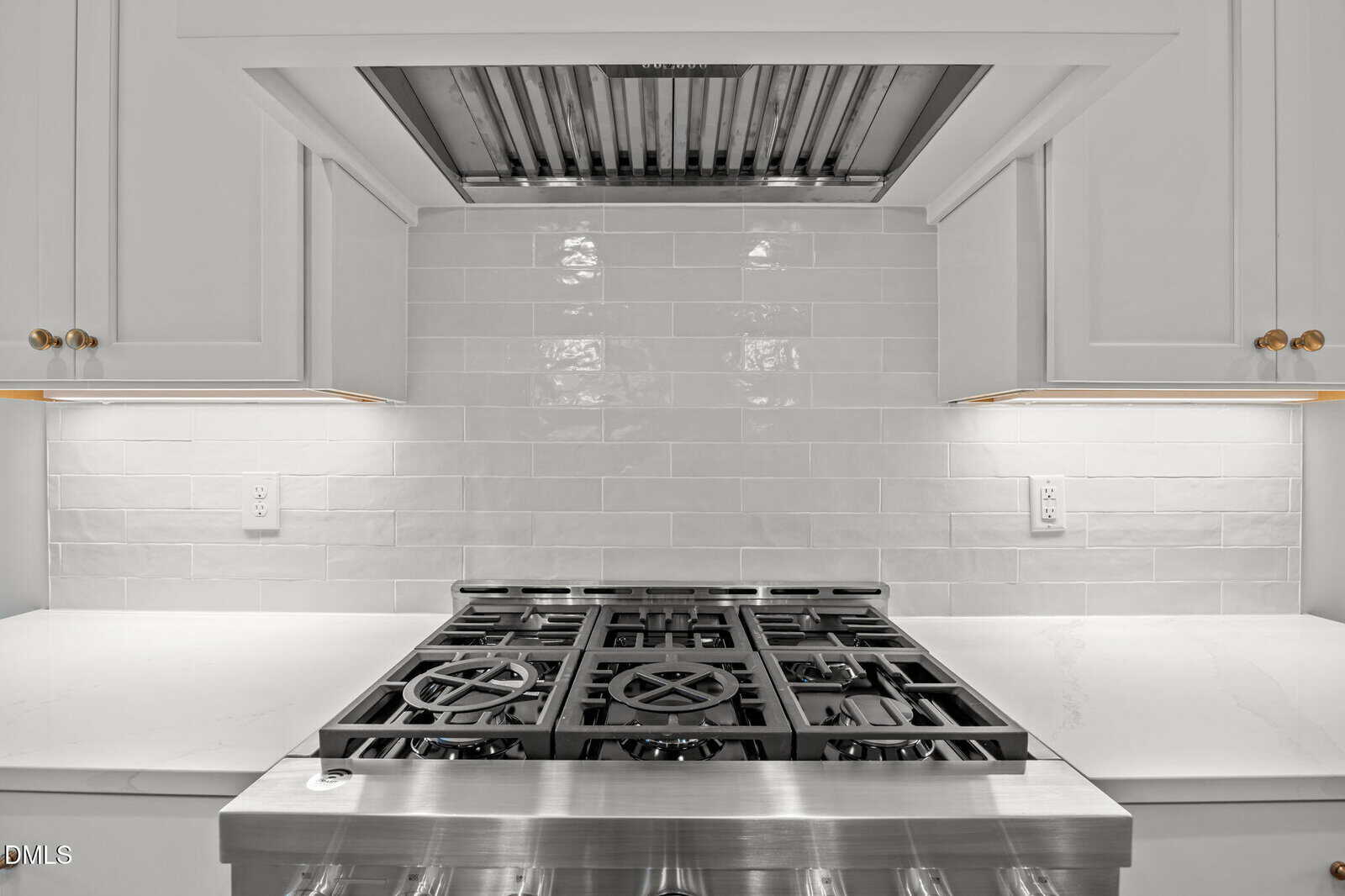 Modern kitchen with a stainless steel gas stove and oven, white subway tile backsplash, white countertops, white cabinets with gold knobs, and a built-in range hood above the stove. The area is brightly lit.