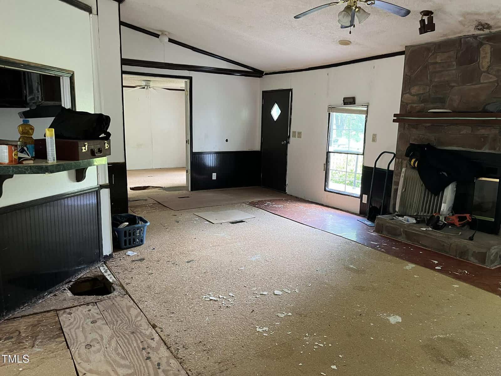 A living room with partially removed flooring, visible subfloor, and scattered debris. There is a stone fireplace, a ceiling fan, and an open doorway to another room. A black door and window let in natural light.