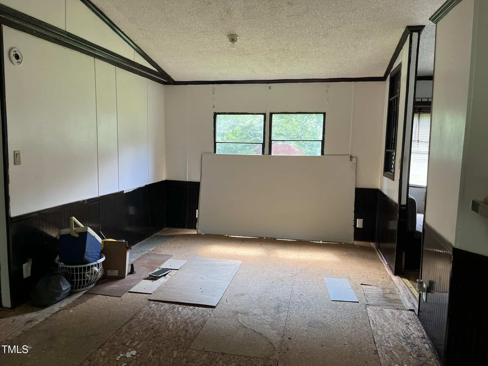 A mostly empty room with white and black walls, a large window at the far end, and a sheet of white material leaning against the window. Boxes and miscellaneous items are in one corner, and the floor is partially unfinished.