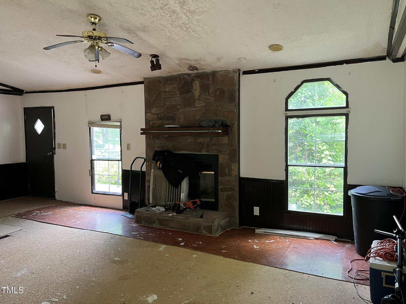 A living room with an unfinished floor, a stone fireplace in the center, a ceiling fan, and two tall windows. There are various items and some debris scattered around the room.