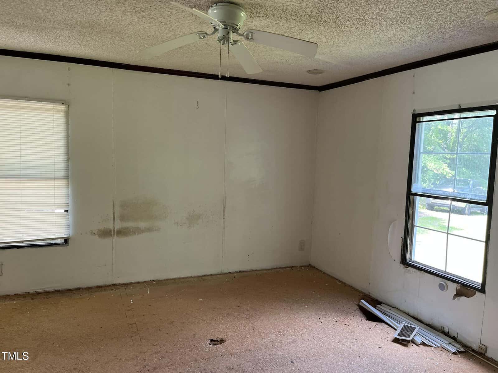 Empty room with bare, brown subfloor, white walls with stains and damage, two windows, a ceiling fan, and scattered debris near a hole in the wall by the right window. Natural light enters from both windows.