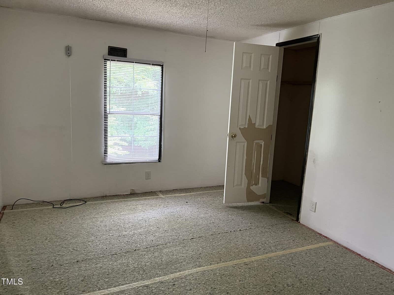 Empty room with a window, open closet door, and unfinished flooring. Light enters through the window, and wires are visible on the floor. The walls are plain white with some paint patches.