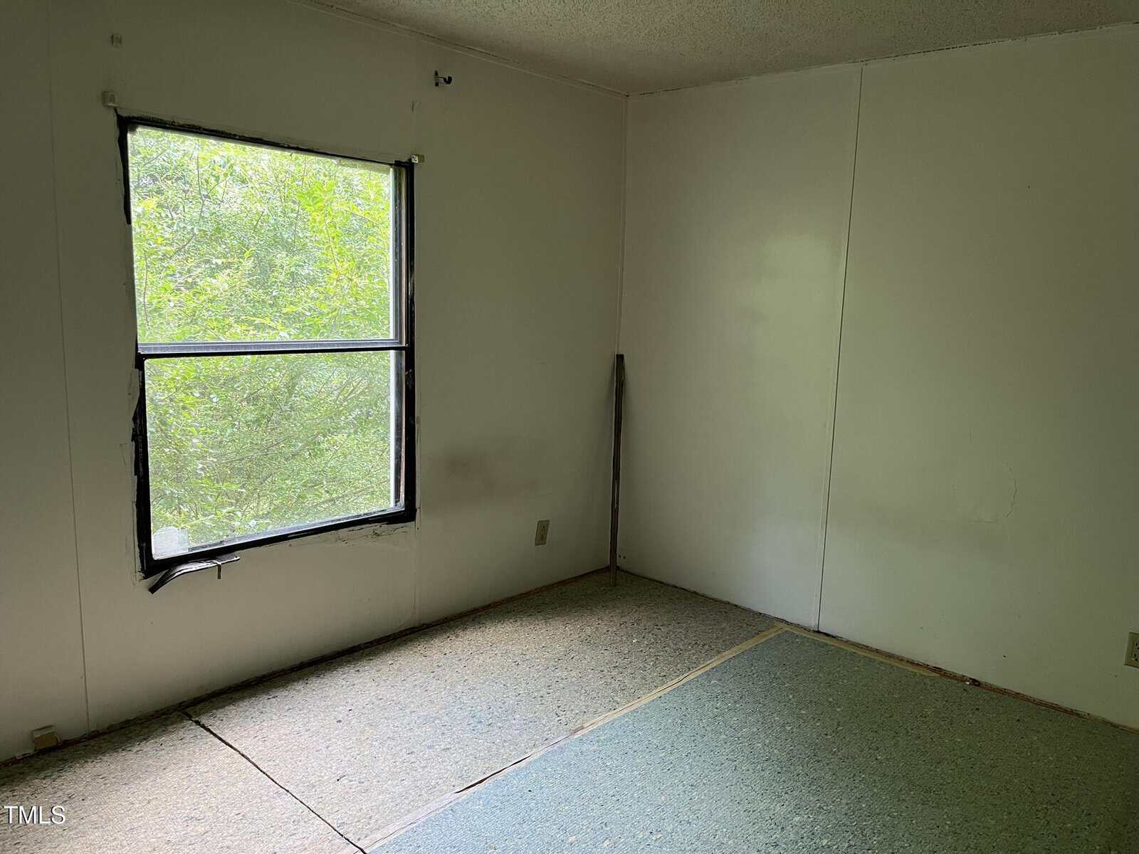 Empty room with white walls, a single window showing greenery outside, and a partially exposed subfloor with some carpet missing.
