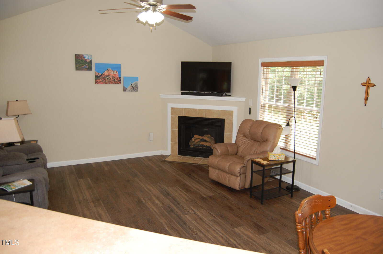 A living room with wood flooring features a fireplace, a mounted TV, an armchair with a side table and lamp, a standing lamp, a window with blinds, wall art, a cross, and part of a dining table and sofa.