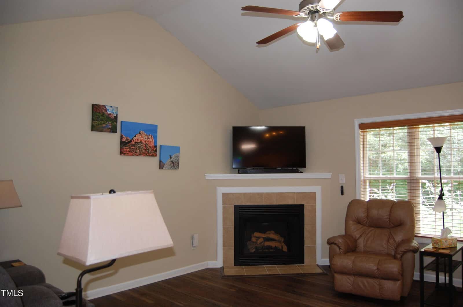 A living room with a beige wall, mounted TV above a fireplace with a tile surround, a brown recliner, a window with blinds, and three small landscape pictures on the wall. A floor lamp and part of a sofa are also visible.