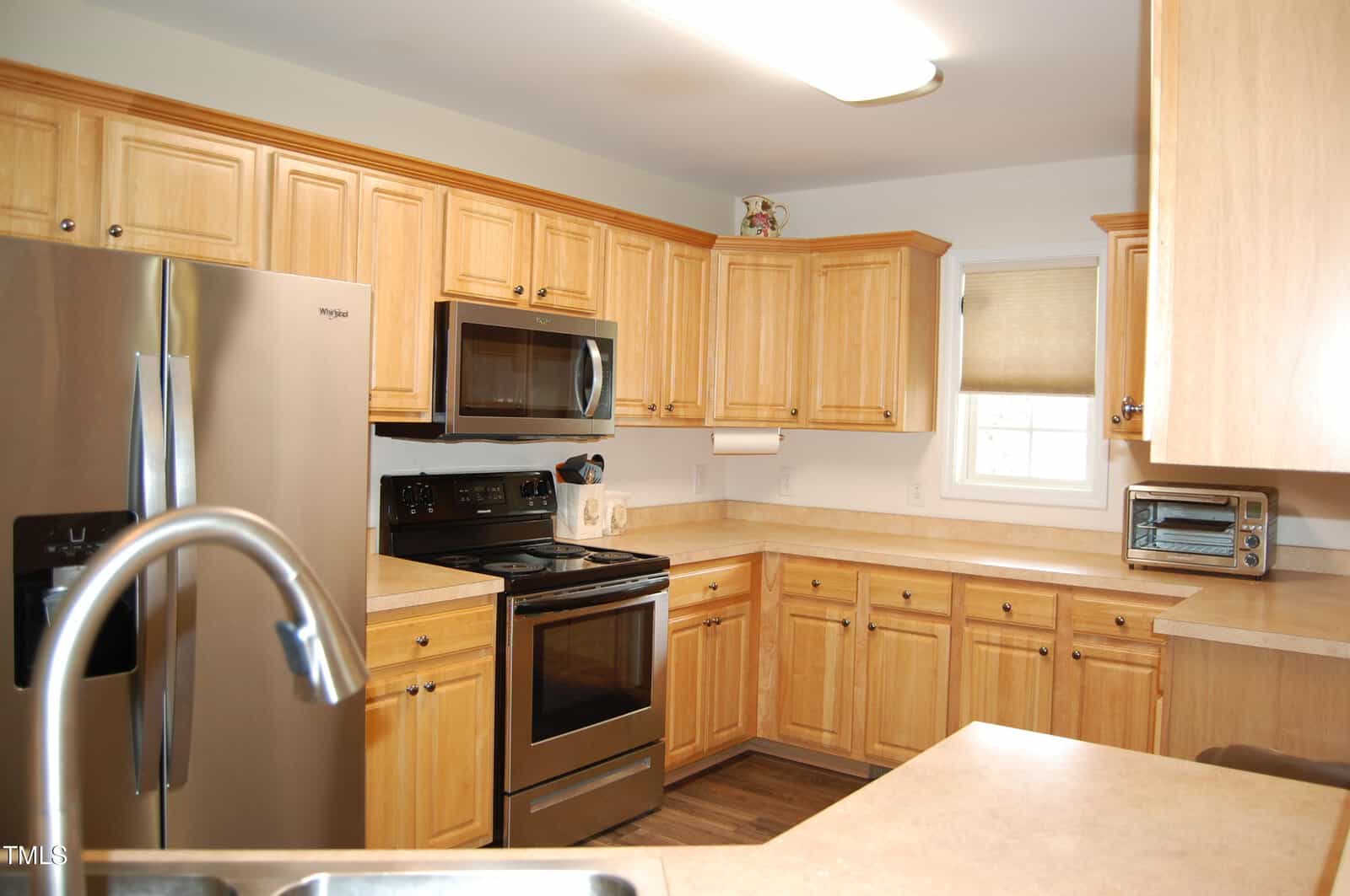 A kitchen with light wood cabinets, stainless steel appliances including a refrigerator and oven, a microwave, a small toaster oven, a sink in the foreground, and a window with a beige shade.