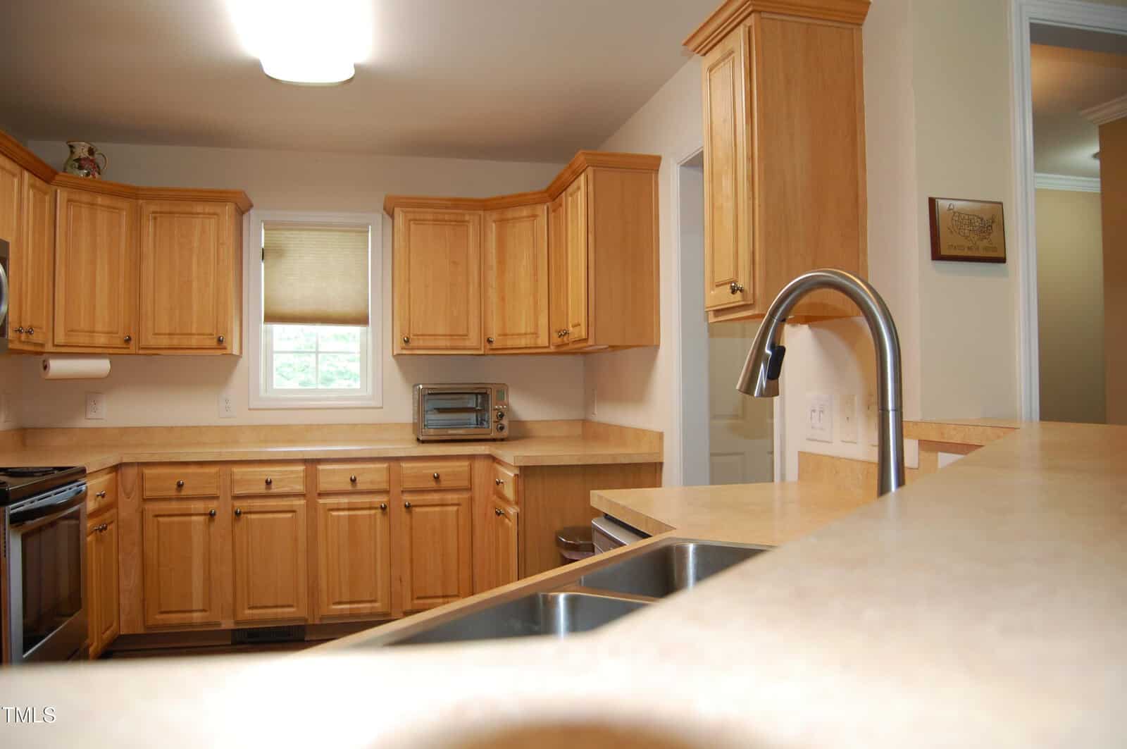 A kitchen with light wood cabinets, beige countertops, a stainless steel sink with a curved faucet, a window with a shade, and a small toaster oven on the counter. The walls are cream-colored.