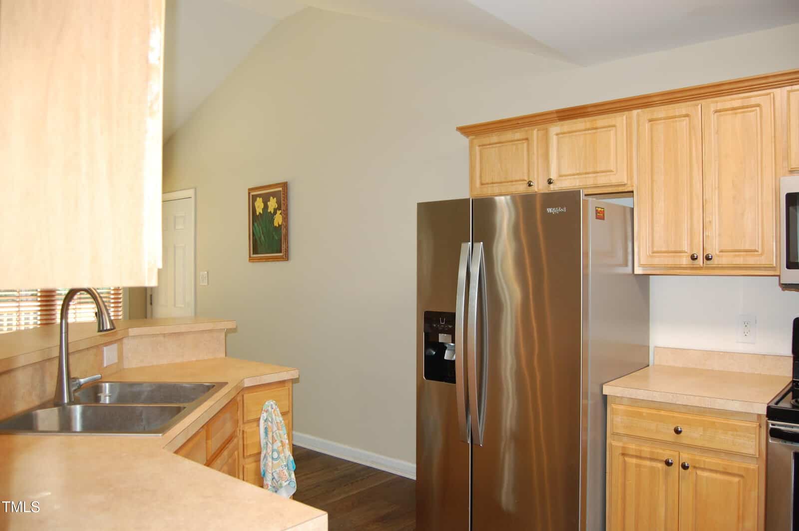 A modern kitchen with light wood cabinets, tan countertops, a stainless steel refrigerator, a double sink, and a wall adorned with a painting of yellow flowers.