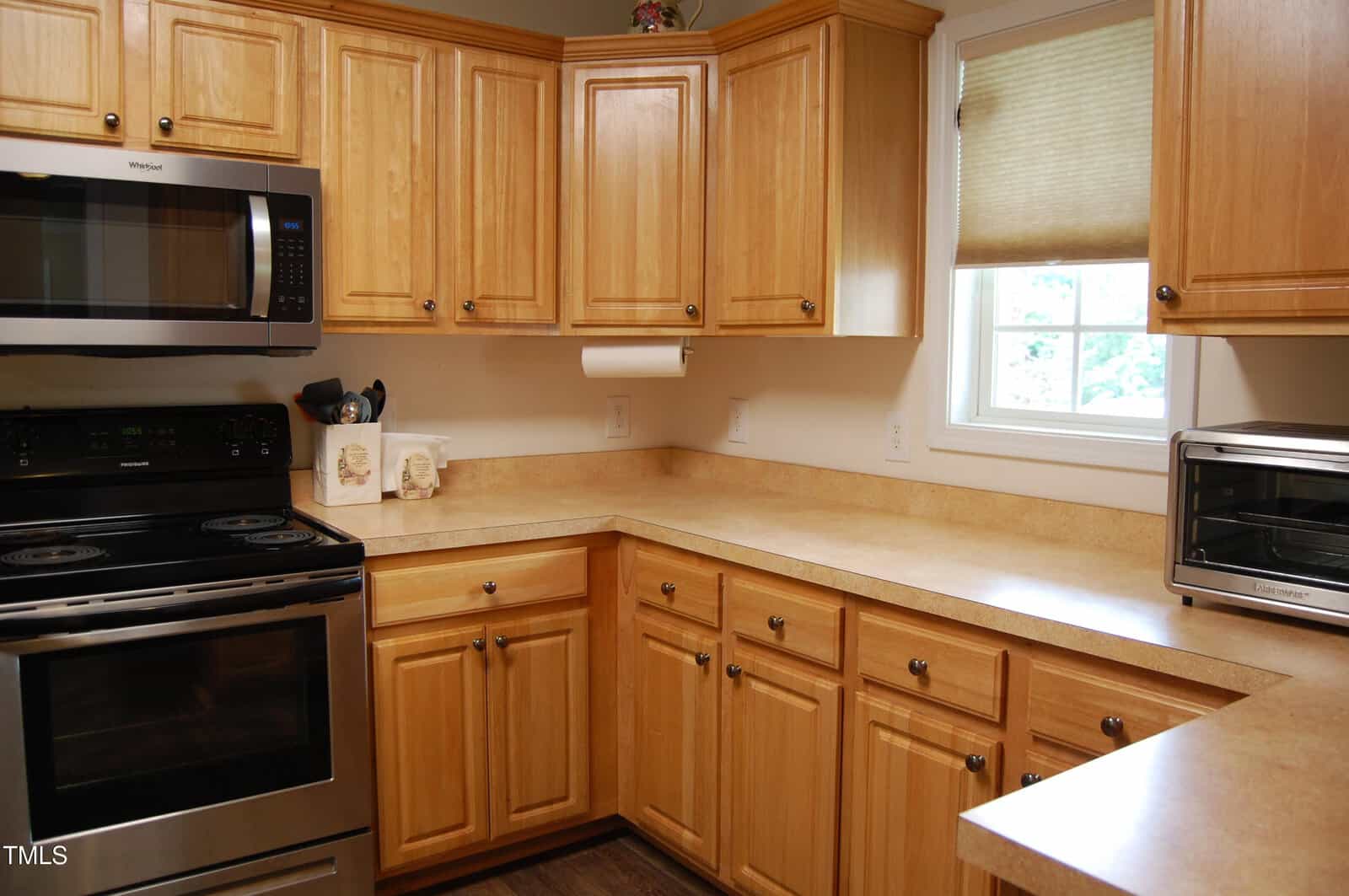 A kitchen with light wood cabinets, beige countertops, a stainless steel oven and microwave, a small toaster oven, and a paper towel holder mounted under the cabinets. A window with a beige shade is above the counter.