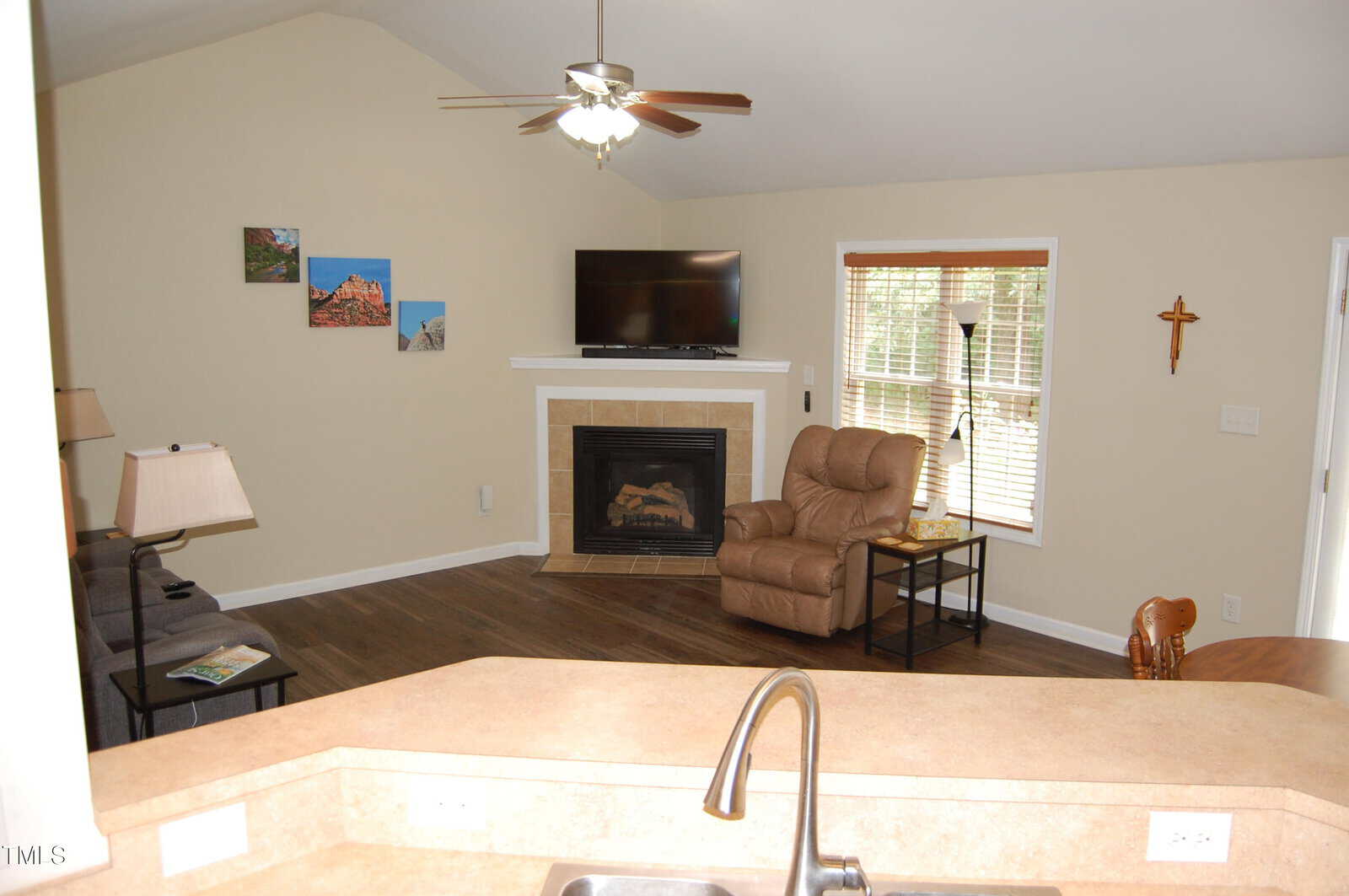 A cozy living room with a recliner, TV above a corner fireplace, three wall art pieces, a side table with a lamp, a ceiling fan, blinds on the window, and a kitchen counter with a sink in the foreground.