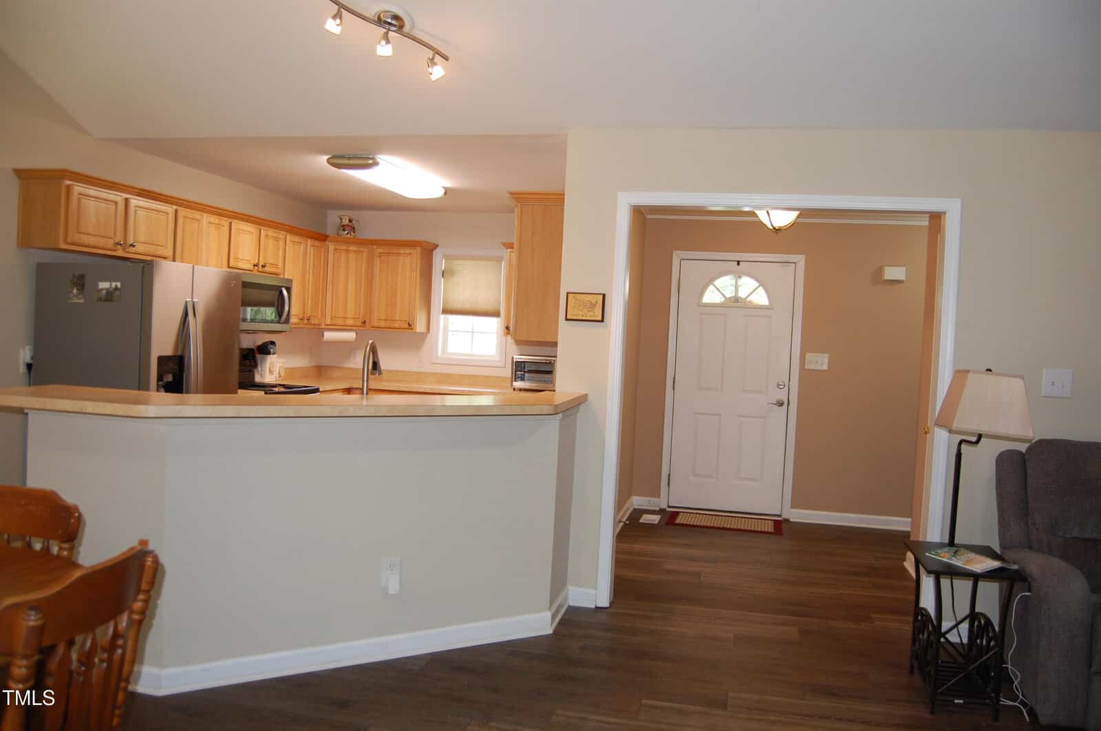 Open kitchen with light wood cabinets and stainless steel appliances, adjacent to a living room area with a white front door, beige walls, hardwood floors, and a small table with a lamp.