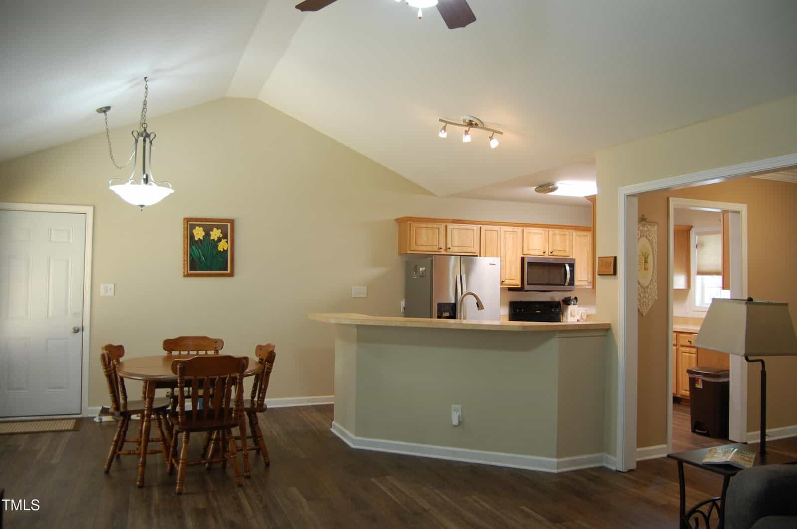 Open kitchen and dining area with wooden table and chairs, light wood cabinets, stainless steel appliances, beige walls, and hardwood floors. A ceiling fan and two pendant lights brighten the space.