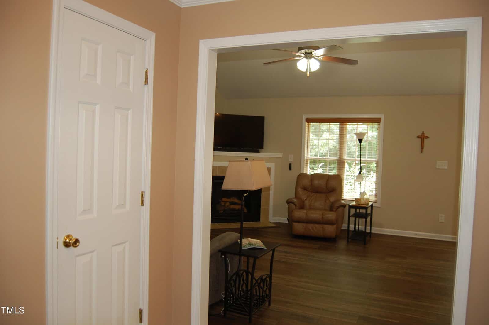 View into a living room with beige walls, a ceiling fan, a brown recliner, a floor lamp, a wall-mounted TV above a fireplace, a window with blinds, and a wooden cross on the wall.