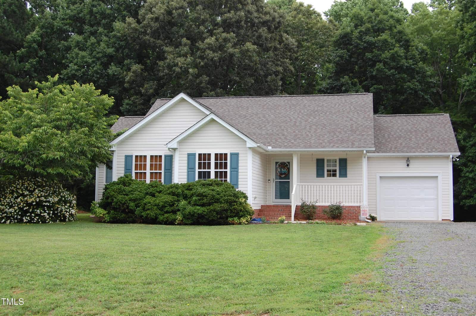 A single-story white house with blue shutters, a front porch, and an attached one-car garage sits on a green lawn with shrubs and trees in the background. The driveway is gravel.