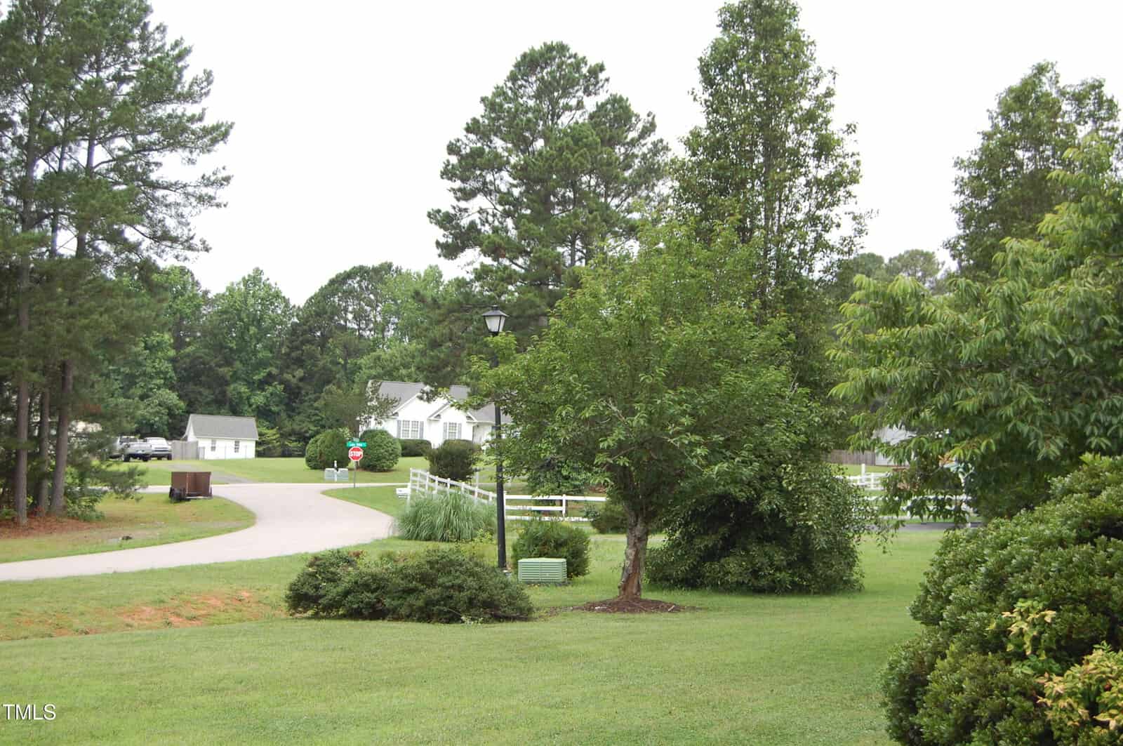 A suburban street curves through a green neighborhood with manicured lawns, trees, bushes, and a lamppost. White houses and a stop sign are visible in the background. The sky is overcast.