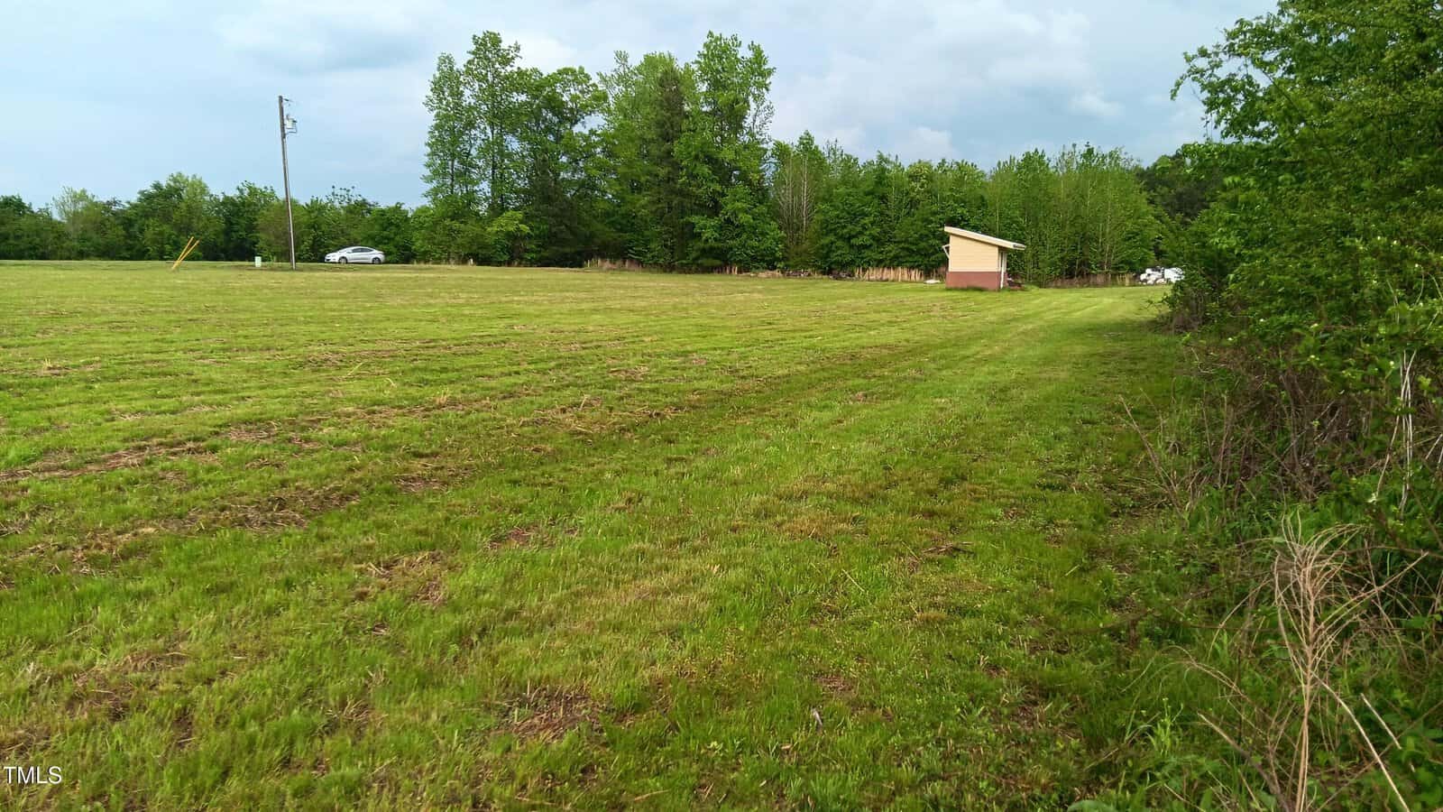 A large grassy field bordered by trees, with a small beige shed near the treeline. Two cars are partially visible in the background along a road running past the field. The sky is mostly cloudy.