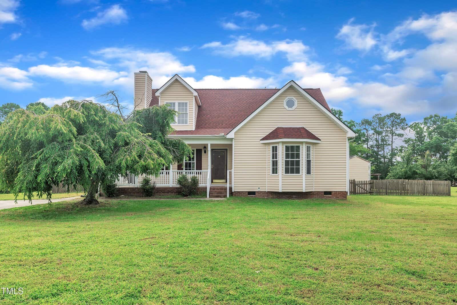 A beige, single-family house with a red roof sits on a large grassy lawn. A tree is on the left, and a wooden fence lines the backyard. The sky is blue with scattered clouds.